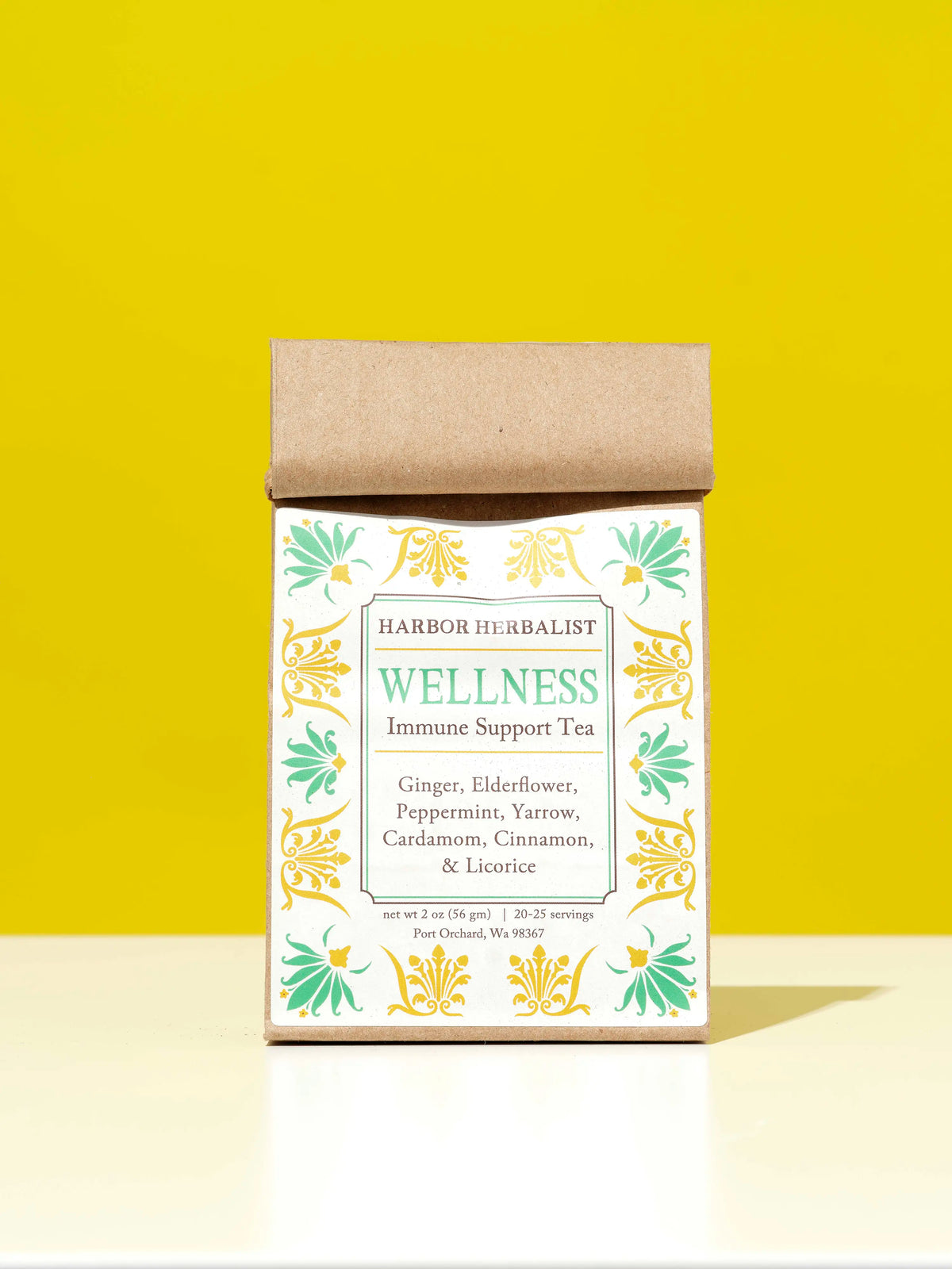 A kraft paper bag of Harbor Herbalist Wellness Immune Support Tea stands on a white surface against a bright yellow background. The packaging features a white label with green and gold botanical illustrations and teal text.