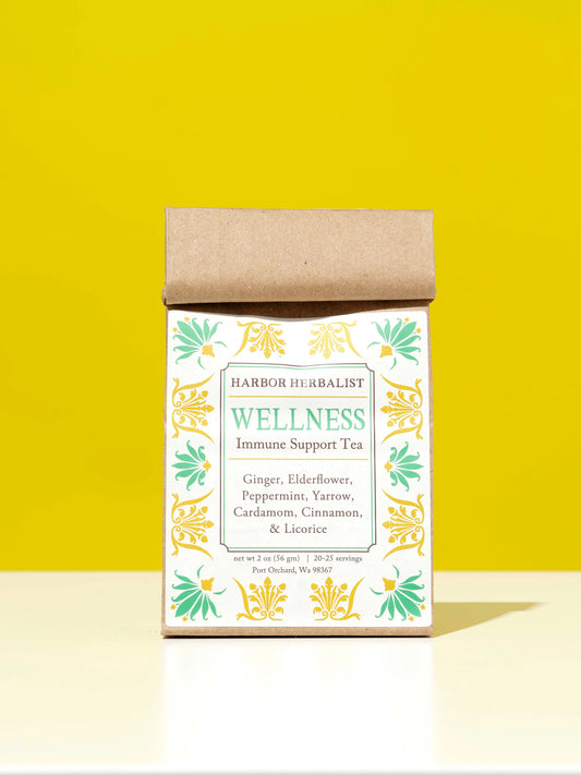 A kraft paper bag of Harbor Herbalist Wellness Immune Support Tea stands on a white surface against a bright yellow background. The packaging features a white label with green and gold botanical illustrations and teal text.