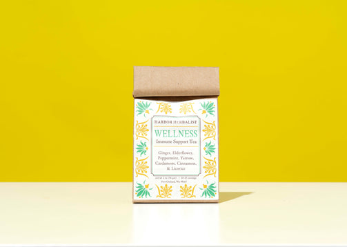A kraft paper bag of Harbor Herbalist Wellness Immune Support Tea stands on a white surface against a bright yellow background. The packaging features a white label with green and gold botanical illustrations and teal text.
