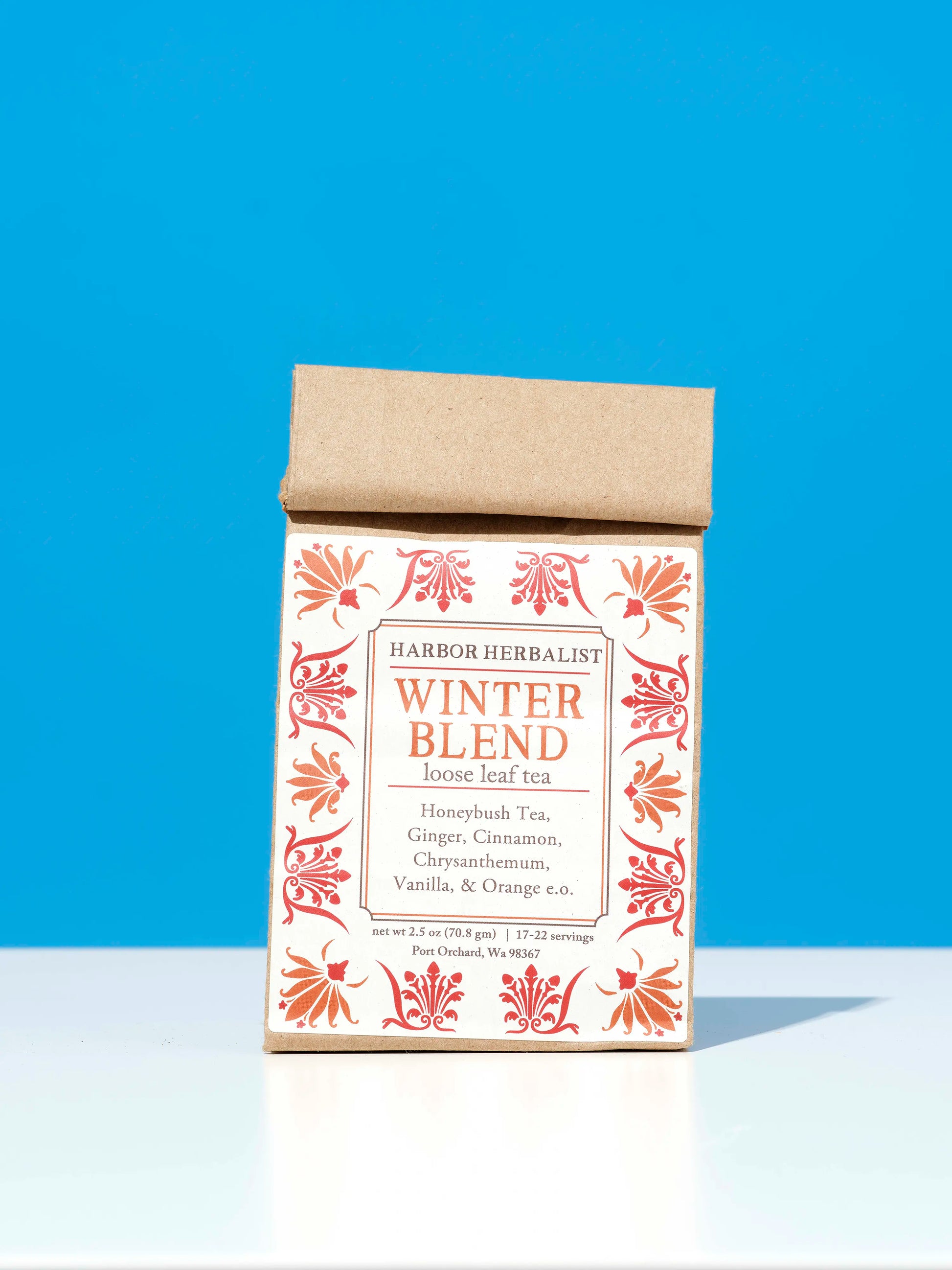 A bag of Harbor Herbalist Winter Blend loose leaf tea, featuring a white label decorated with red and orange botanical illustrations, stands upright on a white surface against a bright blue background.