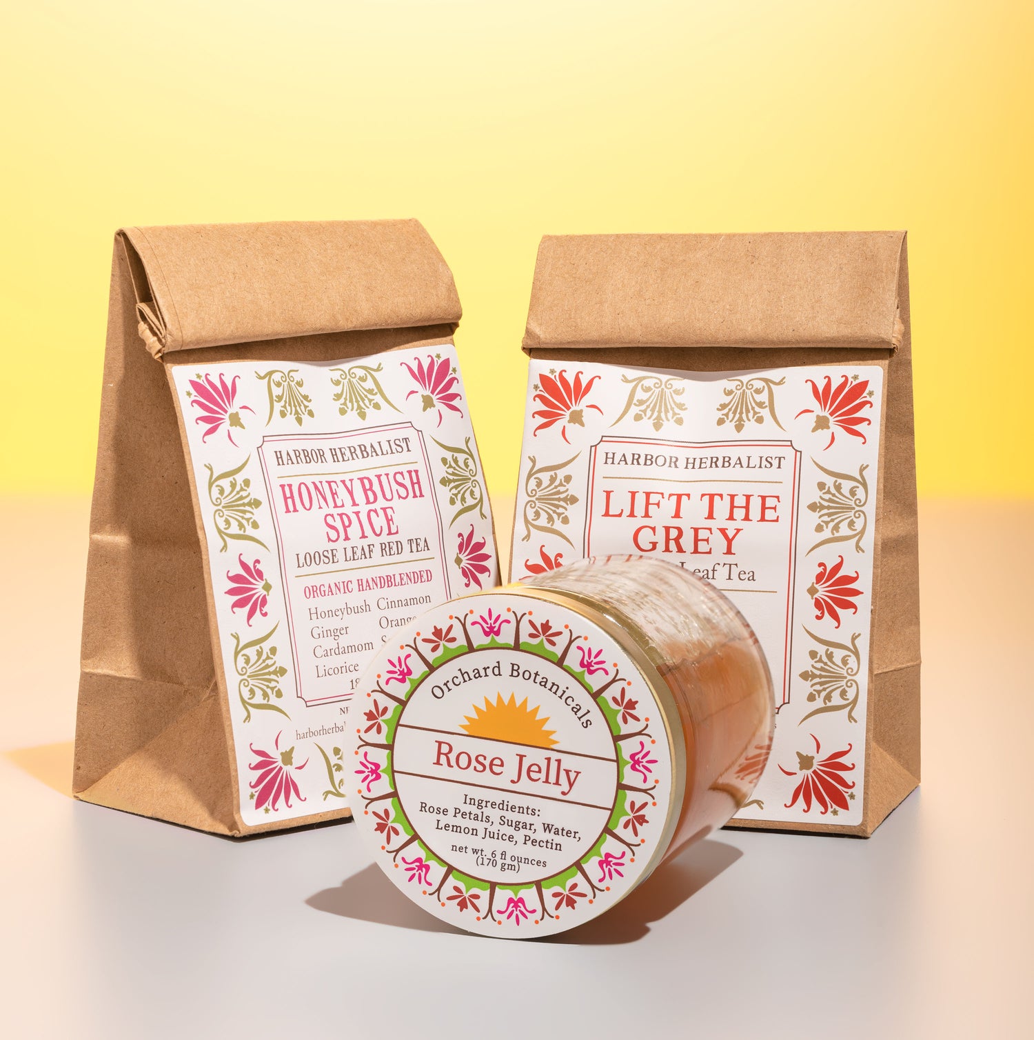 Two natural paper bags of looseleaf tea with the titles "Honeybush Spice" and "Lift The Grey" paired with a tilted glass jam jar titled "Rose Jelly".